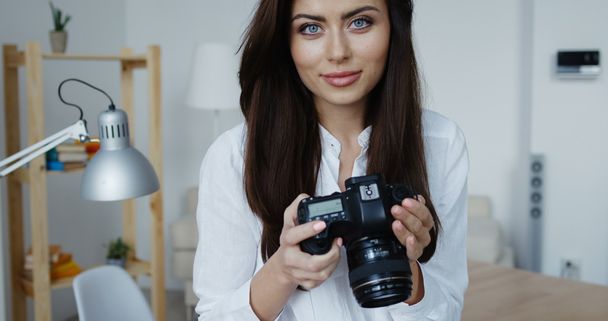 Aantrekkelijke lachende brunette vrouwelijke fotograaf in wit overhemd een bedrijf haar camera, kantoor interieur bij achtergrond afbeelding controleren. - Foto, afbeelding