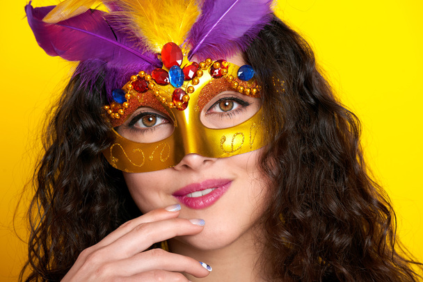 γυναίκα πρόσωπο closeup σε καρναβάλι μάσκα μεταμφίεση με φτερό, όμορφο κορίτσι πορτρέτο σε κίτρινο χρώμα φόντο, μακριά σγουρά μαλλιά - Φωτογραφία, εικόνα