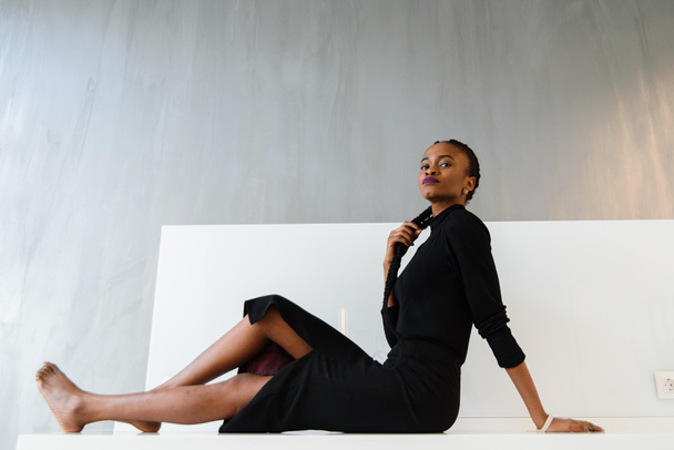 Elegant african or black american woman in dark dress posing on desk in light interior - Fotografie, Obrázek