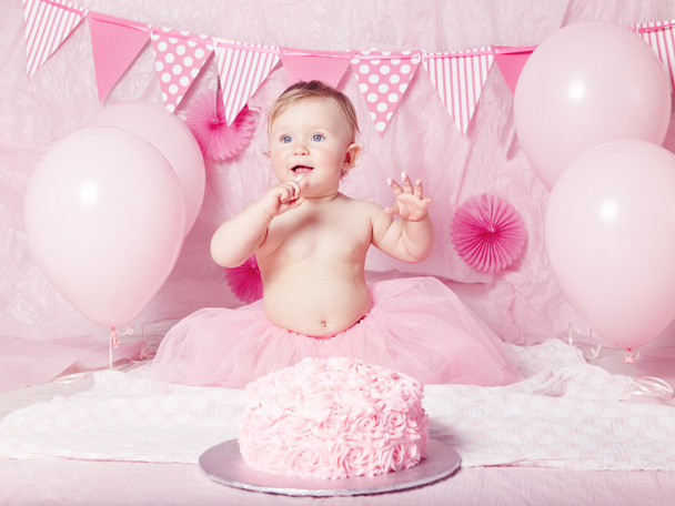 Portrait of cute adorable Caucasian baby girl with blue eyes in pink tutu skirt celebrating her first birthday with gourmet cake and balloons looking away, cake smash first year concept - Fotó, kép
