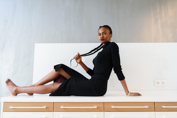 Elegant african or black american woman in dark dress posing on desk in light interior - Foto, imagen