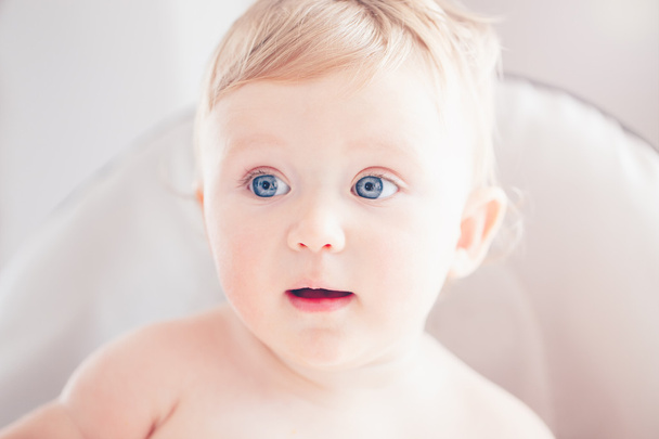 Primer plano retrato de lindo adorable divertido rubia caucásica sonriente bebé niña con ojos azules con expresión de cara emocional sentado en la silla alta en la cocina mirando hacia otro lado
 - Foto, Imagen