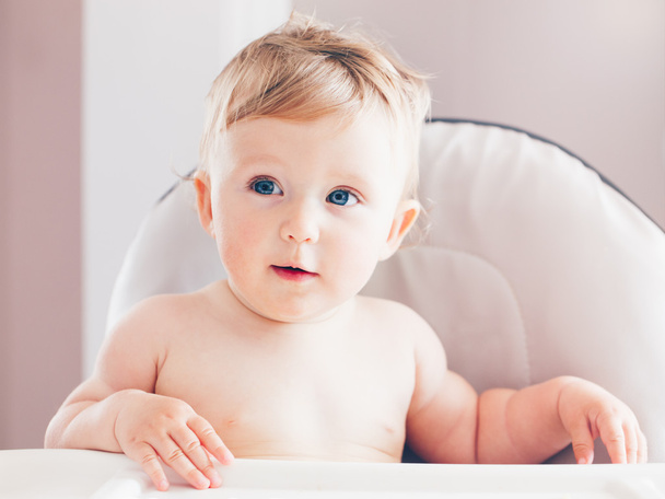 Gros plan portrait de mignon adorable drôle blonde caucasien souriant bébé garçon fille aux yeux bleus avec expression émotionnelle visage assis dans la chaise haute dans la cuisine détournant les yeux
 - Photo, image