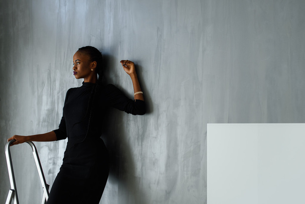 Beautiful and sexy black woman in dark dress posing looking away leaning hand on gray wall at studio - Фото, зображення
