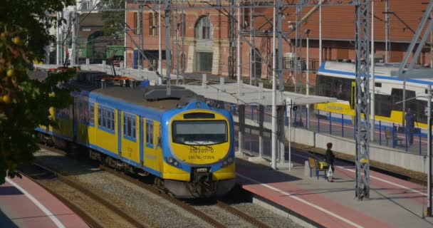 People at the Platphorm Are Waiting Blue and Yellow Passenger Electric Train is Standing at the Railway Station Another Train Arrives Buildings Roofs - Materiaali, video