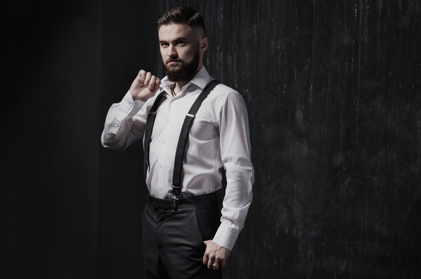 Homem barbudo atraente em uma camisa branca e suspensórios de pé perto da parede escura
. - Foto, Imagem