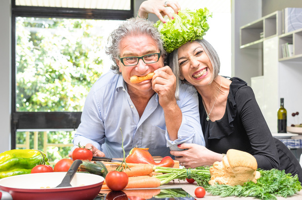 Senior couple having fun in kitchen with healthy food - Retired people cooking meal at home with man and woman preparing lunch with bio vegetables - Happy elderly concept with mature funny pensioner - Foto, Bild