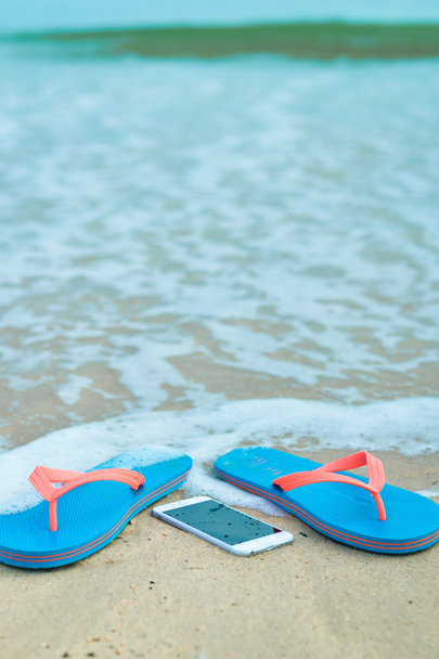 Top view close up on mobile phone with slippers on summer beach background. Happy joyful vacation - Foto, afbeelding