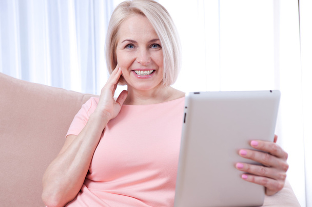 Smiling beautiful middle-aged woman sitting on couch with a tabl - Foto, Bild