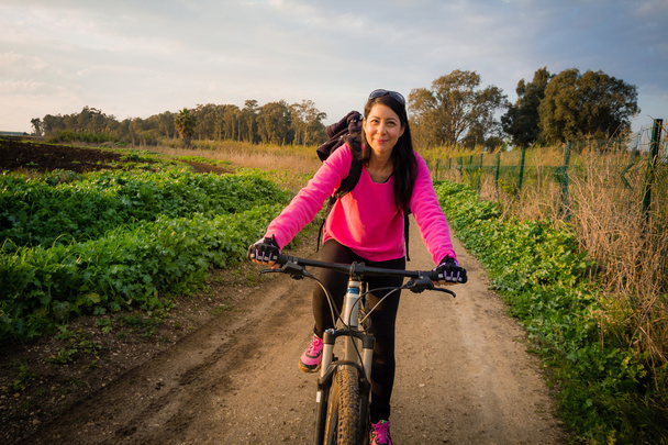 Жінка їде на велосипеді в сільській місцевості і дивиться на камеру
 - Фото, зображення