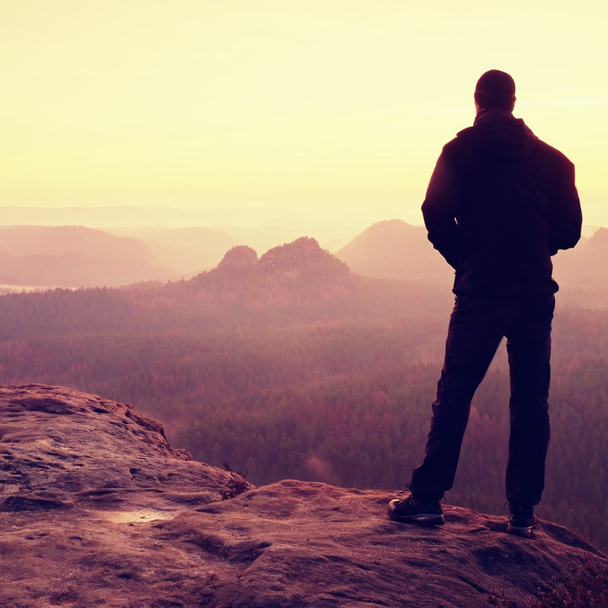 Turista com mochila e postes no pico rochoso olhando para o sol. amanhecer ensolarado nas montanhas, nebuloso vale sonhador abaixo
. - Foto, Imagem