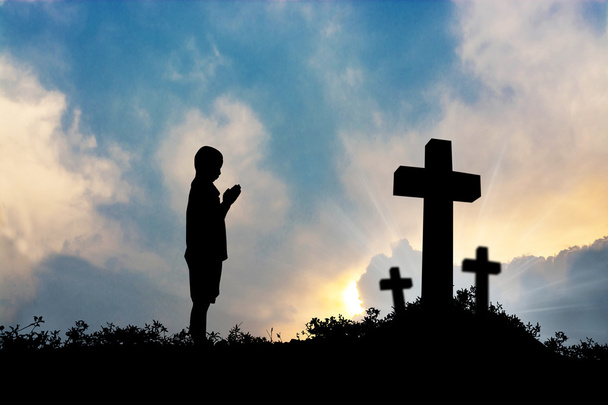 Silhueta de menino orando a uma cruz com céu nebuloso conceito de pôr do sol para a religião, adoração, amor e espiritualidade
 - Foto, Imagem