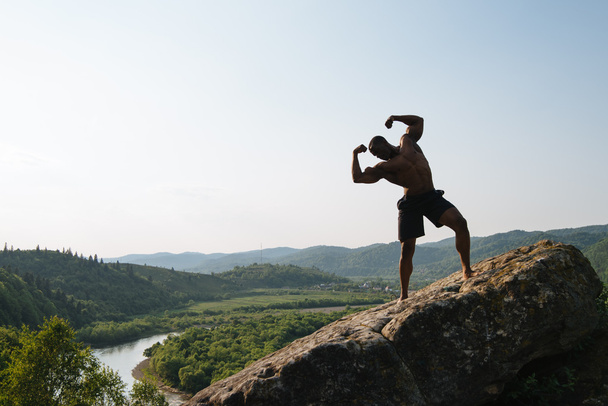 Silhouette de bodybuilder afro-américain avec torse nu posant sur le rocher. Montagne verte nature fond
 - Photo, image