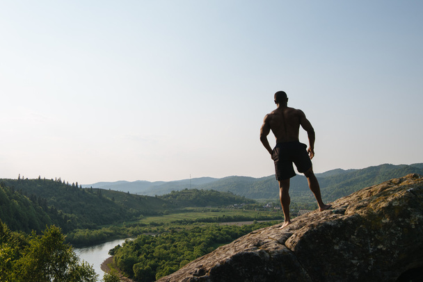 Σιλουέτα αφροαμερικάνων αθλητικό άνδρα με γυμνό τον κορμό που παρουσιάζουν στο βράχο. Φύση φόντο πράσινο βουνό - Φωτογραφία, εικόνα