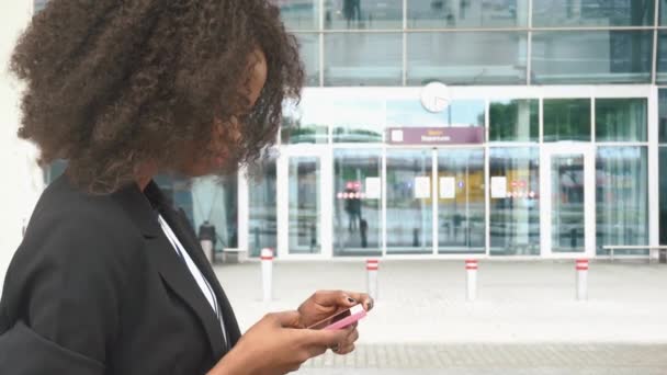 Side view of beautiful african american business lady texting on phone while waiting for a flight in airport. Urban glass wall on background - Felvétel, videó