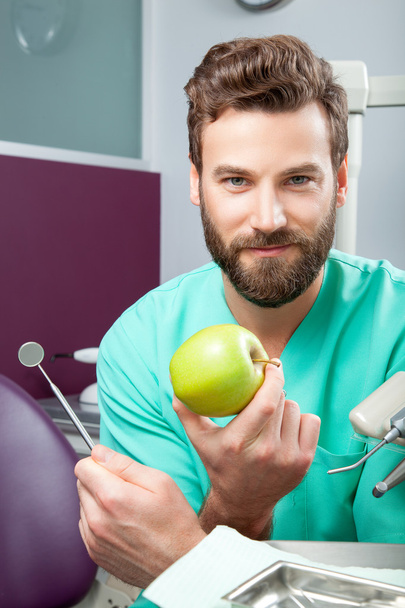 Молодой красивый мужчина-врач улыбается белыми зубами, держа яблоко
 - Фото, изображение