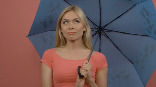 Dreaming blondynka z tajemniczym uśmiechem patrząc od gospodarstwa niebieski parasol, na białym tle na różowym tle - Materiał filmowy, wideo