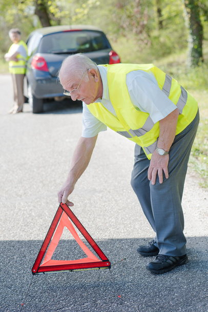 Elderly couple broken down in car, man putting red warning triangle on road - Photo, image