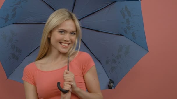 Attractive smiling caucasian blonde woman playing with blue umbrella on pink background - Πλάνα, βίντεο