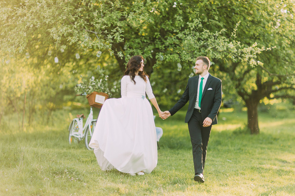 Šťastná novomanželená nevěsta a ženich chodící po trávě v parku se svým bicyklem poblíž stromu na pozadí - Fotografie, Obrázek