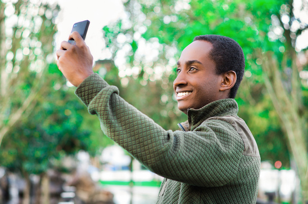 Handsome hispanic black man wearing green sweater in outdoors park area holding up phone and watching screen as in taking selfie - Фото, изображение