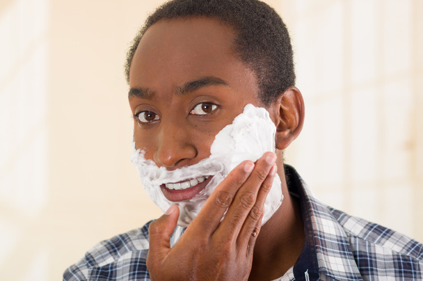 Young man wearing white blue square pattern shirt applying shaving foam onto face using hands, looking into camera - Foto, afbeelding