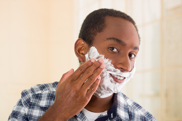 Young man wearing white blue square pattern shirt applying shaving foam onto face using hands, looking into camera - 写真・画像