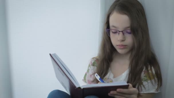 Serious little girl in glasses diligently does his homework at window. - Video