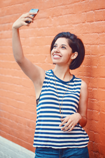 Porträt der schönen jungen lateinamerikanischen hispanischen Mädchen Frau in gestreiftem T-Shirt und blauen Jeans mit dunklem kurzen langen Bob Frisur außerhalb in der Nähe der roten Backsteinmauer in der Stadt macht Selfie-Foto mit Handy - Foto, Bild
