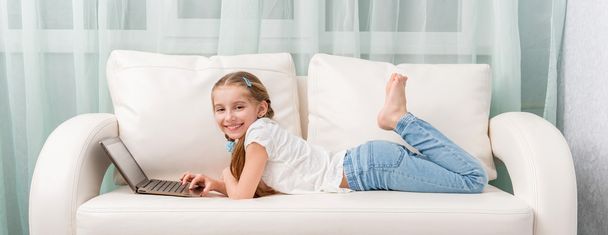 little girl lying on sofa looks at notebook and laughs - Photo, Image