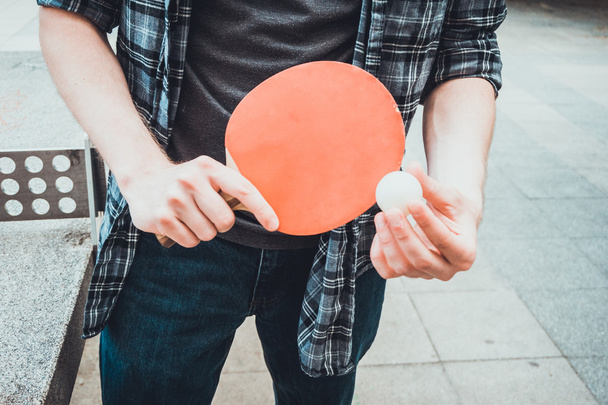 junger Mann mit einem bunten roten Tischtennis- oder Tischtennisschläger und -ball neben einem Allwettertisch im Freien, die Hände in Großaufnahme - Foto, Bild
