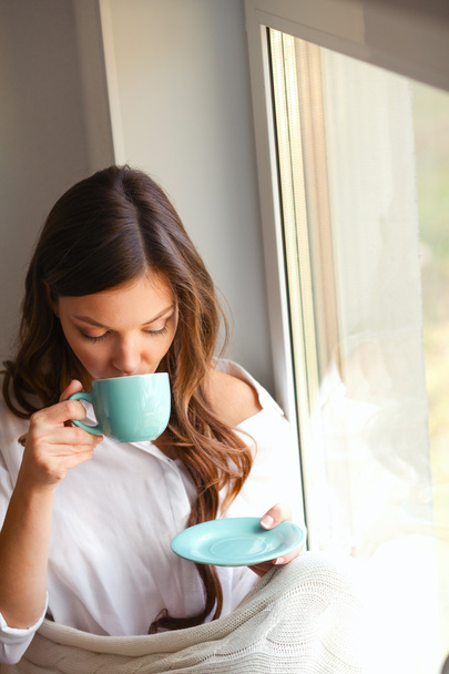 Νεαρή γυναίκα στο σπίτι κάθεται κοντά στο παράθυρο χαλαρώνοντας στο σαλόνι της διαβάζοντας βιβλίο και πίνοντας καφέ ή τσάι - Φωτογραφία, εικόνα