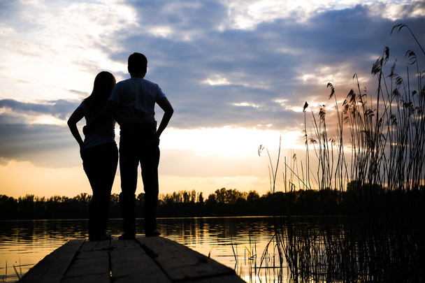 Ζευγάρι στέκεται στη γέφυρα στην όχθη της λίμνης στο ηλιοβασίλεμα - Φωτογραφία, εικόνα
