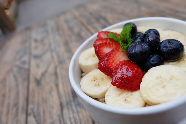Acai bowl with fresh fruit strawberry, blueberry, banana and peppermint leaves on top on the wooden table. - Photo, Image