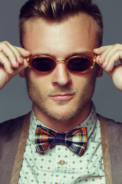 Emotive portrait of handsome young man in retro shirt, scottish in cell bow-tie and round unusual sunglasses posing over gray background. White shiny smile and healthy skin. Urban style. Studio shot - Photo, Image