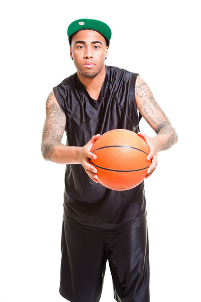 Retrato de estudio del jugador de baloncesto con gorra verde de pie y sosteniendo la pelota aislada en blanco. Tatuajes en sus brazos
. - Foto, Imagen