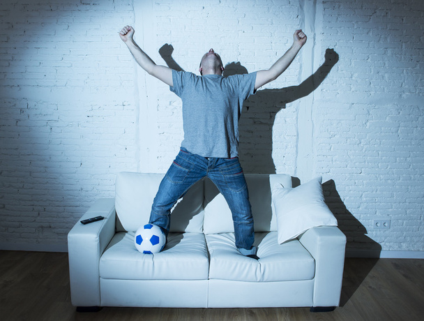 fanatic and crazy football fan watching television soccer match with ball jumping on sofa celebrating goal - Photo, Image