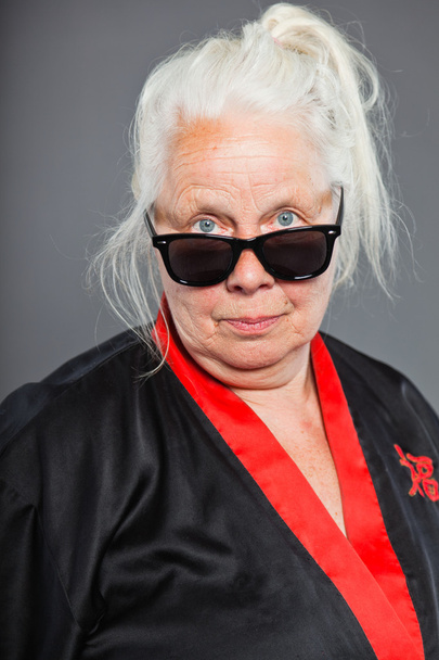 Старшая женщина с длинными седыми волосами в черном и красном кимоно и черных солнцезащитных очках. Круто выглядишь. Съемка изолирована на сером фоне
. - Фото, изображение