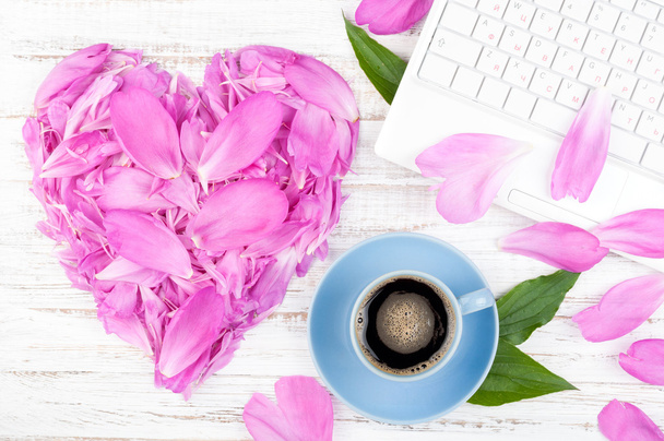 Werkruimte met laptop, kopje koffie en bloemen op houten achtergrond. Pioenrozen bloemblaadjes in hart vorm - Foto, afbeelding