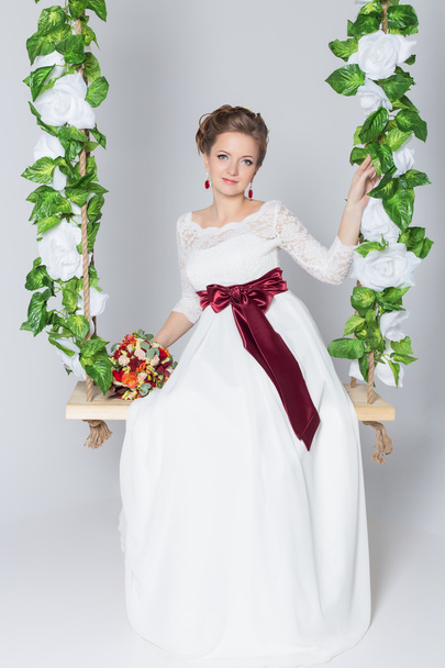 beautiful lovely bride is sitting on a swing with a beautiful bouquet of colorful flowers in a white dress with evening hairstyle - Foto, imagen