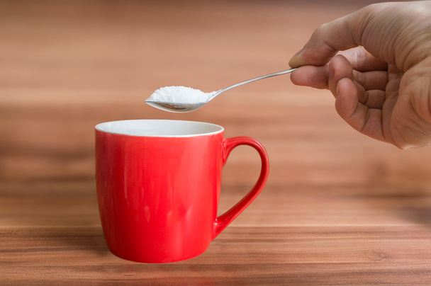 Hand holds teaspoon with sugar above red cup of tea. Unhealthy eating concept. - Foto, imagen