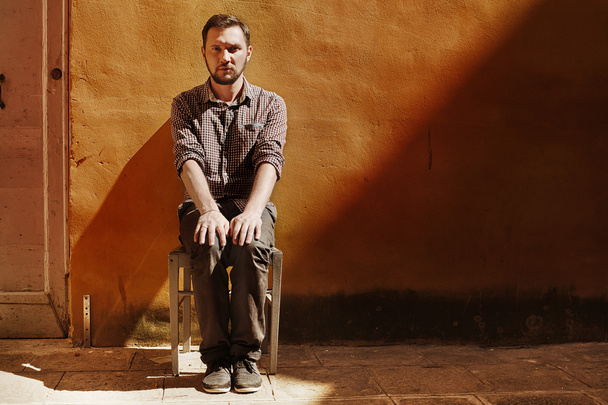 White male sitting on a wooden table on an empty street at the house with yellow walls - Fotó, kép