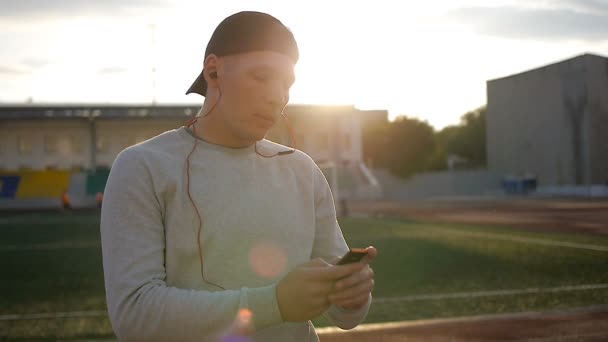 Slowmotion of young man wears headphones on field at sunset on stadium track - Metraje, vídeo