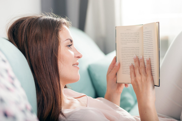 Young woman lying on couch and reading a book at home. Casual style indoor shoot - 写真・画像