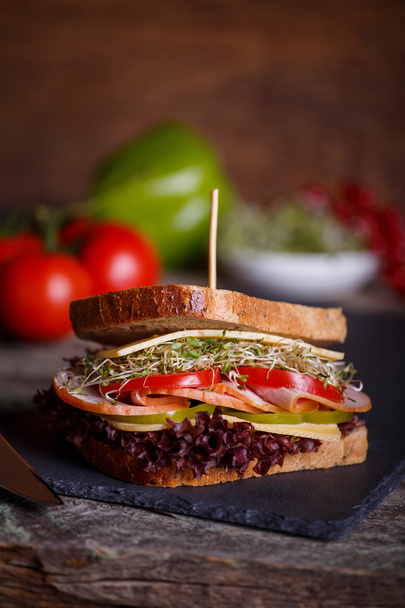 Sandwich with cereals bread with lettuce, slices of fresh tomatoes and sweet pepper, ham, prosciutto, salami, cheese  and  onion sprouts on dark marble background. Close-up - Foto, Bild