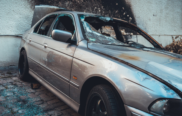 Úplně zpopelněný vyhořelý luxusní sedan na spáleném černém dlážděném nádvoří, pohled zblízka rozbitými okny zčernalého interiéru vozidla - Fotografie, Obrázek