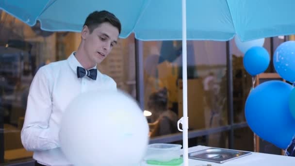 Junger Kerl, der Zuckerwatte an einer speziellen Maschine herstellt, trägt Fliege, hinter ihm Luftballons - Filmmaterial, Video