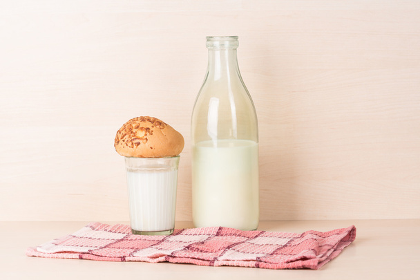facettiertes Glas Milch mit einem Brötchen darauf steht auf einem roten quadratischen Küchentuch neben einer traditionell geöffneten altmodischen Flasche Milch. - Foto, Bild