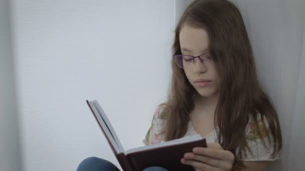Serious little girl in glasses diligently does his homework at window. - Metraje, vídeo