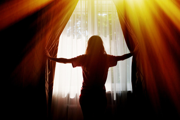 Silhouet van een jonge vrouw opent gordijnen bij raam - Foto, afbeelding